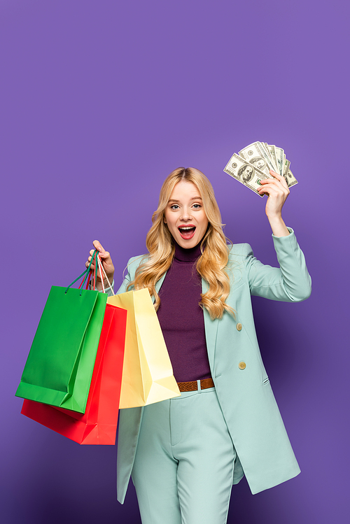 happy blonde young woman in fashionable turquoise blazer with shopping bags and money on purple background