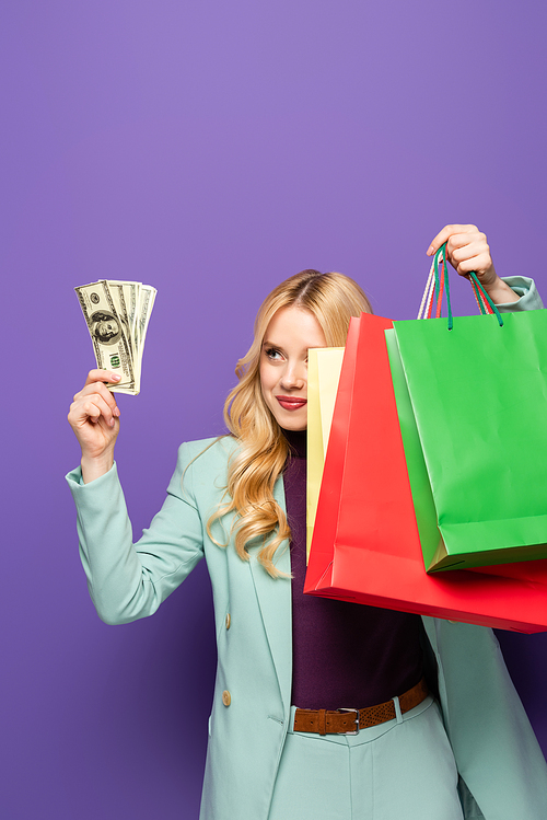 blonde young woman in fashionable turquoise blazer with shopping bags and money on purple background