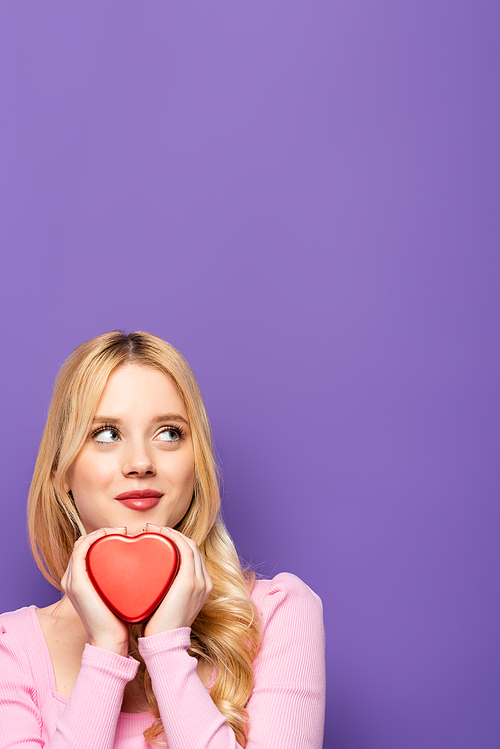 dreamy blonde young woman holding red heart shaped box on purple background
