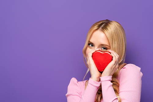 blonde young woman holding red heart shaped box near face on purple background