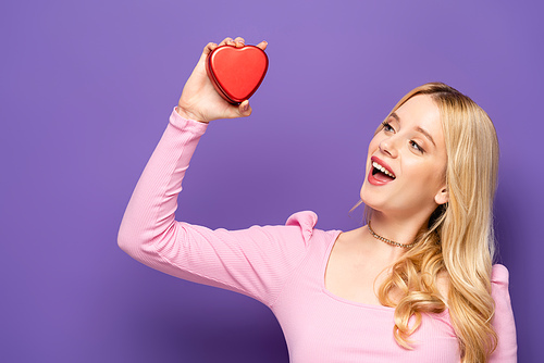 happy blonde young woman holding red heart shaped box on purple background