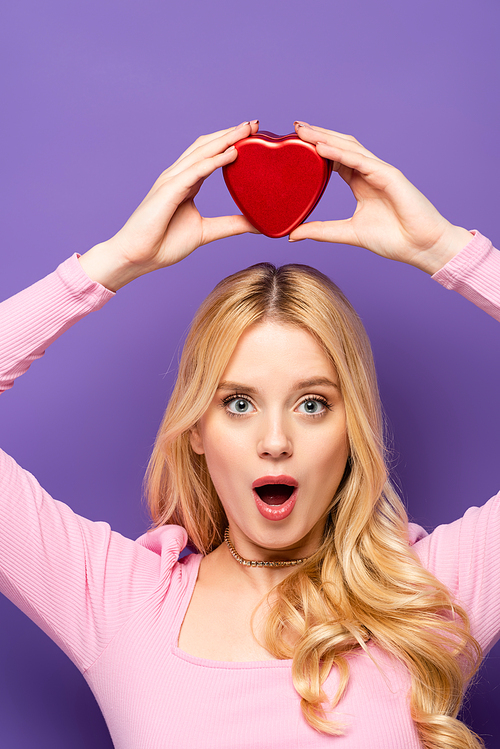 shocked blonde young woman holding red heart shaped box above head on purple background
