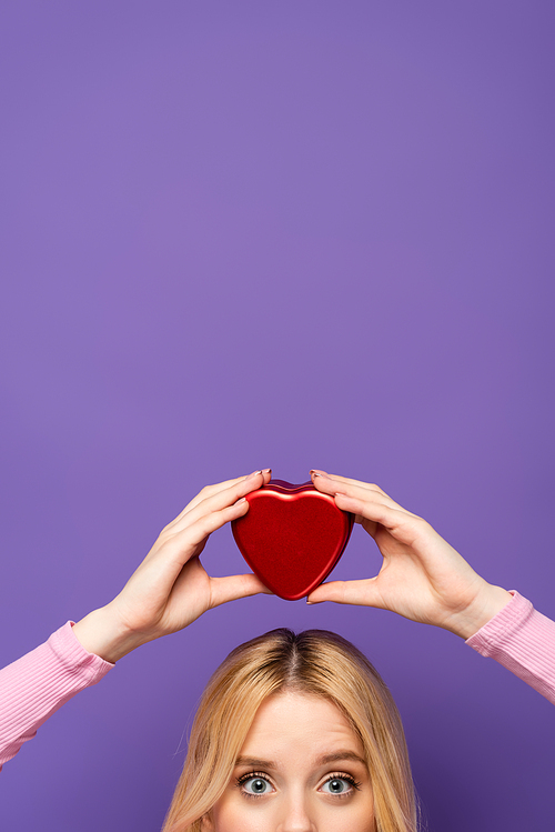 cropped view of blonde young woman holding red heart shaped box on head on purple background