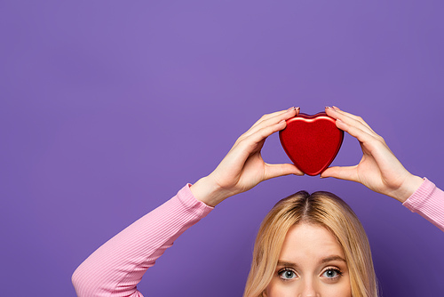 cropped view of blonde young woman holding red heart shaped box on head on purple background