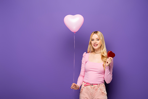 smiling blonde young woman holding heart shaped balloon and gift on purple background