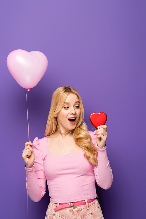 happy blonde young woman holding red heart shaped box and balloon on purple background