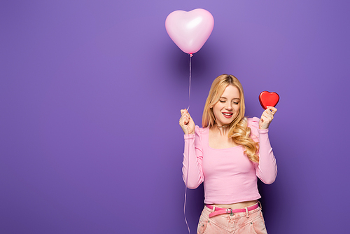 happy blonde young woman holding red heart shaped box and balloon on purple background