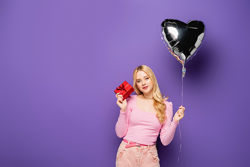 blonde young woman holding heart shaped balloon and gift on purple background