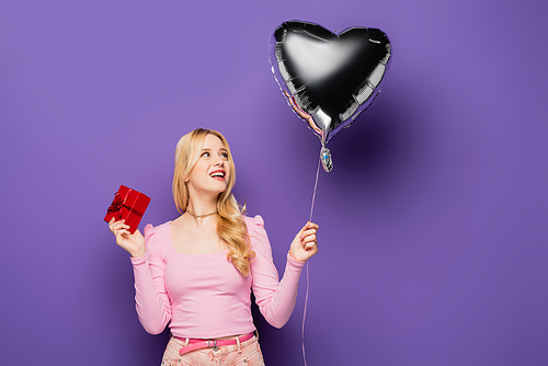 happy blonde young woman holding heart shaped balloon and gift on purple background