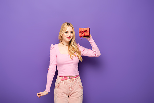 happy blonde young woman holding red gift box on purple background