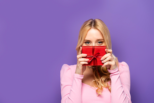 blonde young woman holding red gift box near face on purple background