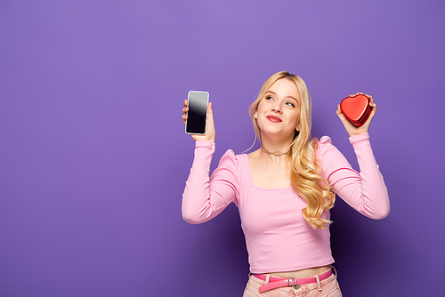 blonde young woman with red heart shaped box and smartphone on purple background