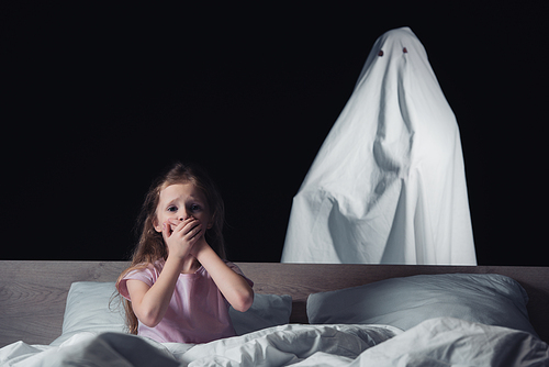 frightened child sitting in bed and showing hush sign while white ghost standing behind bed isolated on black