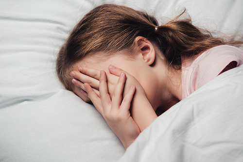 scared child covering face with hands while lying on white bedding