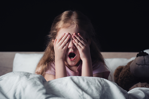 frightened kid screaming and covering eyes with hands while sitting on bedding near teddy bear isolated on black