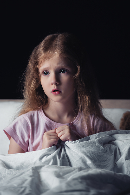 frightened kid looking away while sitting on bedding isolated on black