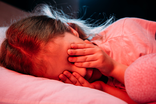 scared child covering eyes with hands while lying in bed isolated on black