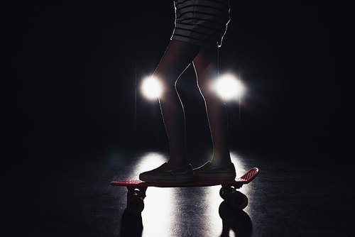 cropped view of kid riding penny board in darkness with illumination of headlights on black background