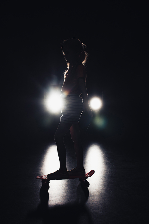 kid riding penny board in darkness with illumination of headlights on black background