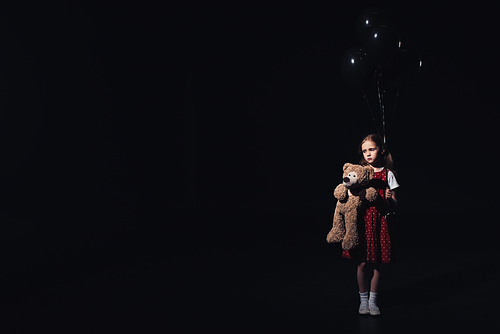 depressed, lonely child holding black balloons and teddy bear isolated on black