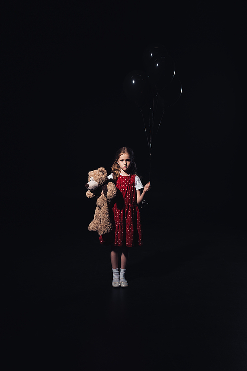 offended kid holding black balloons and teddy bear while  isolated on black