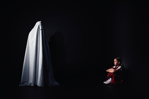 scared child sitting on floor and looking at terrible white ghost on black background