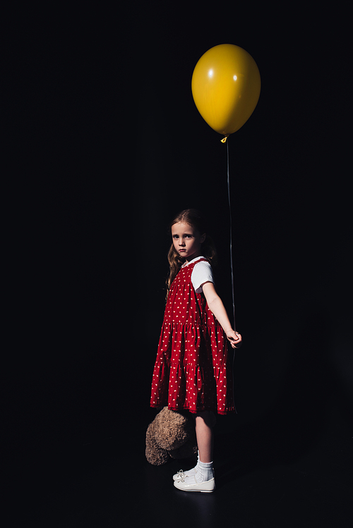 offended child holding yellow balloon and teddy bear while  isolated on black