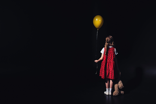 back view of depressed child with yellow balloon and teddy bear isolated on black