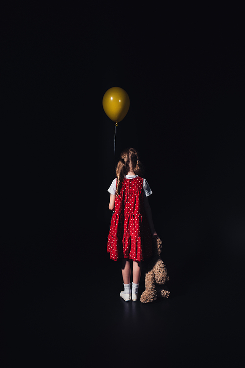 back view of lonely child with yellow balloon and teddy bear isolated on black
