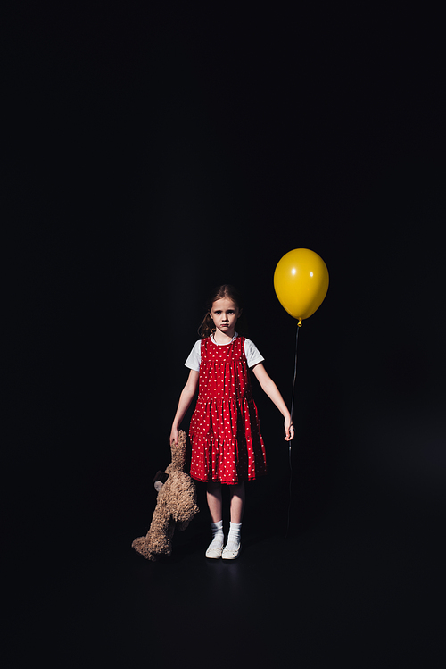 upset, lonely kid with yellow balloon and teddy bear  isolated on black