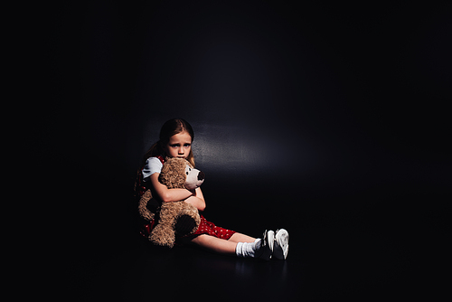 lonely, scared kid sitting on floor and hugging teddy bear on black background