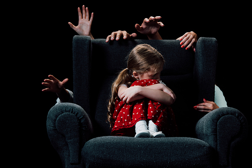 scared child sitting in armchair surrounded with human hands isolated on black