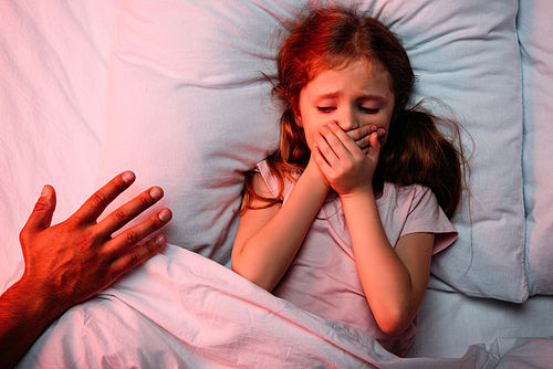 male hand near frightened child lying in bed and showing hush sign