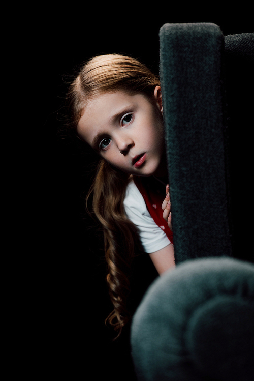 anxious child  while hiding behind armchair isolated on black