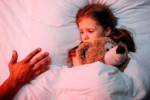 male hand near scared child lying in bed with teddy bear