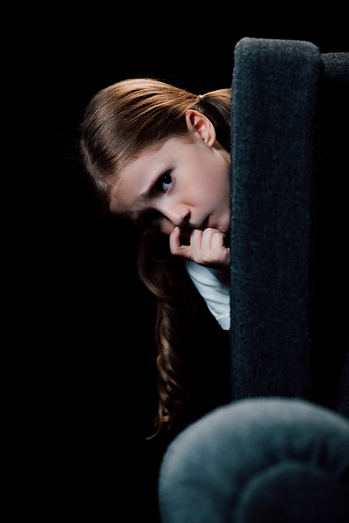 frightened child  while hiding behind armchair isolated on black