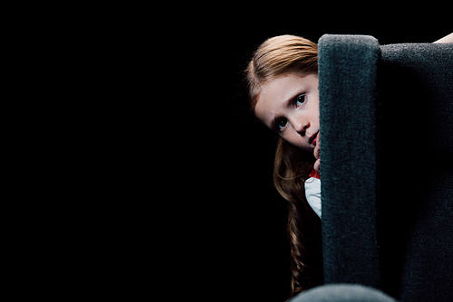 scared child  while hiding behind armchair isolated on black