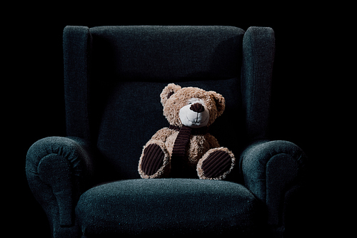 beige teddy bear in grey soft armchair isolated on black