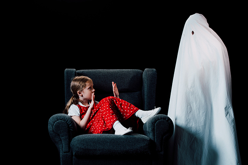 direful ghost standing near scared girl sitting in armchair isolated on black