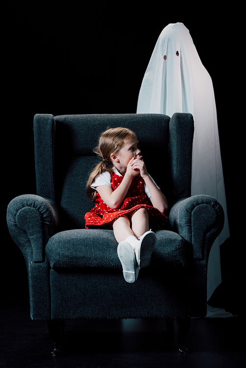 frightened child showing hush sign near white ghost standing behind armchair isolated on black
