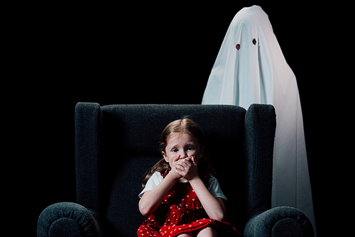 white ghost and scared child sitting in armchair and showing hish sign isolated on black