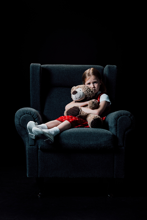 depressed, scared child sitting in armchair and hugging teddy bear isolated on black