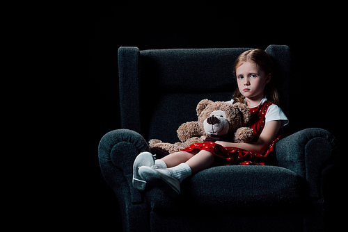 sad, lonely child sitting in armchair and holding teddy bear isolated on black