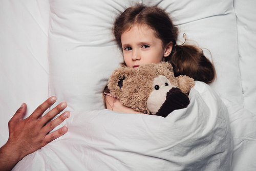 cropped view of male hand near scared child lying in bed with teddy bear