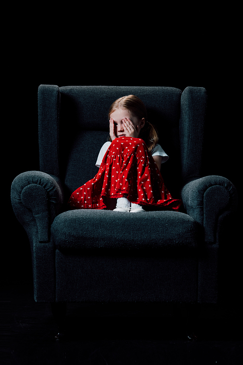 frightened, depressed child sitting in armchair and covering eyes with hands isolated on black