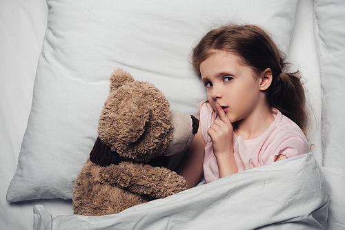 top view of scared child showing hush sign and  while lying in bed with teddy bear