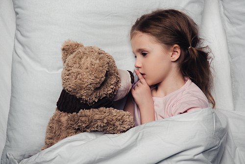 adorable child showing hush sign while lying in bed with teddy bear