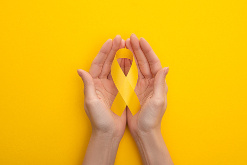 Partial view of female hands with yellow awareness ribbon on colorful background, international childhood cancer day concept