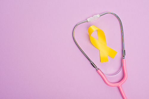 Top view of yellow ribbon and pink stethoscope on violet background, international childhood cancer day concept