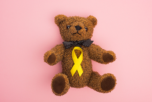 Top view of yellow awareness ribbon on teddy bear with bow on pink background, international childhood cancer day concept